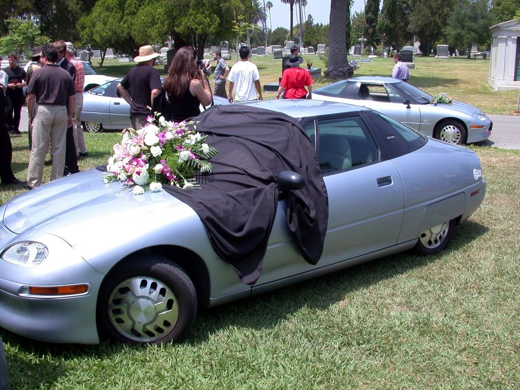 A GM EV1 draped in black cloth with flowers on its bonnet at a mock funeral in 2003